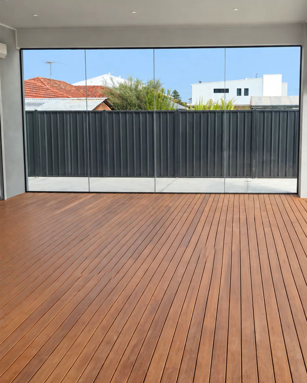 A wall of four frameless sliding glass panels closing an alfresco on a timber deck, with the fence and neighbouring roofs visible through the glass