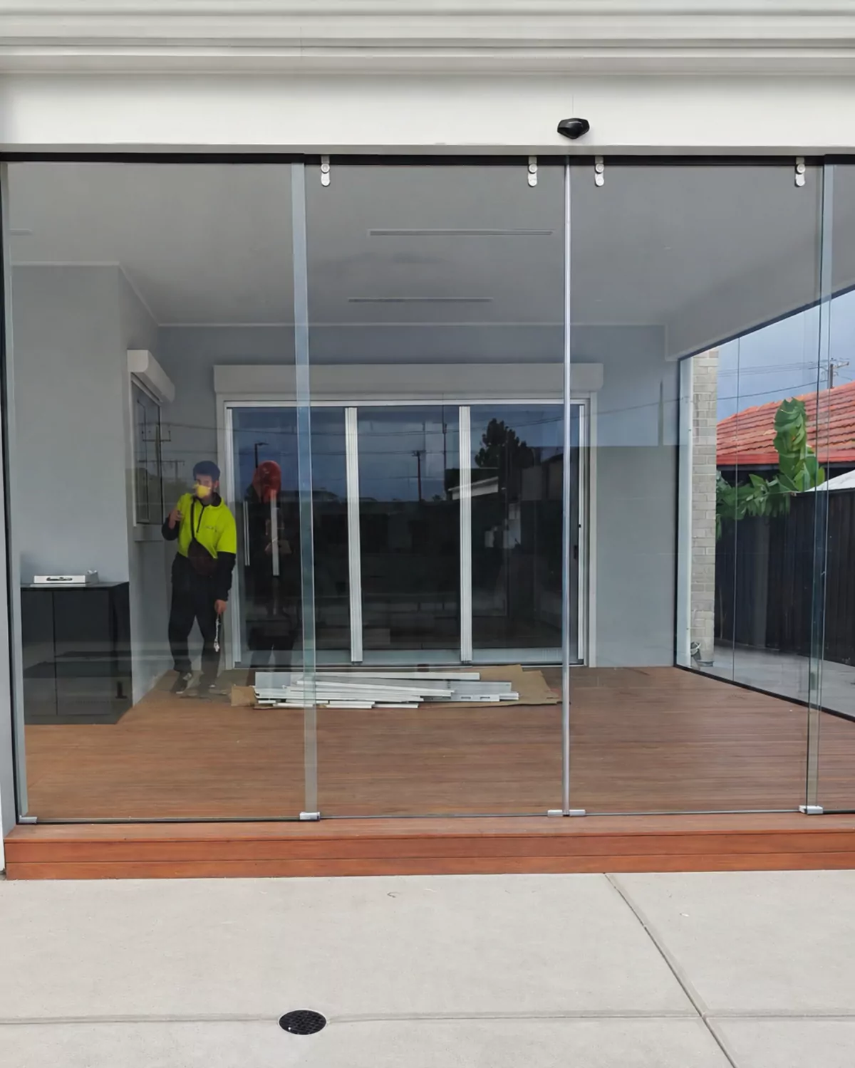 Frameless glass panels hung from a top track across a timber-decked alfresco, with an installer inside