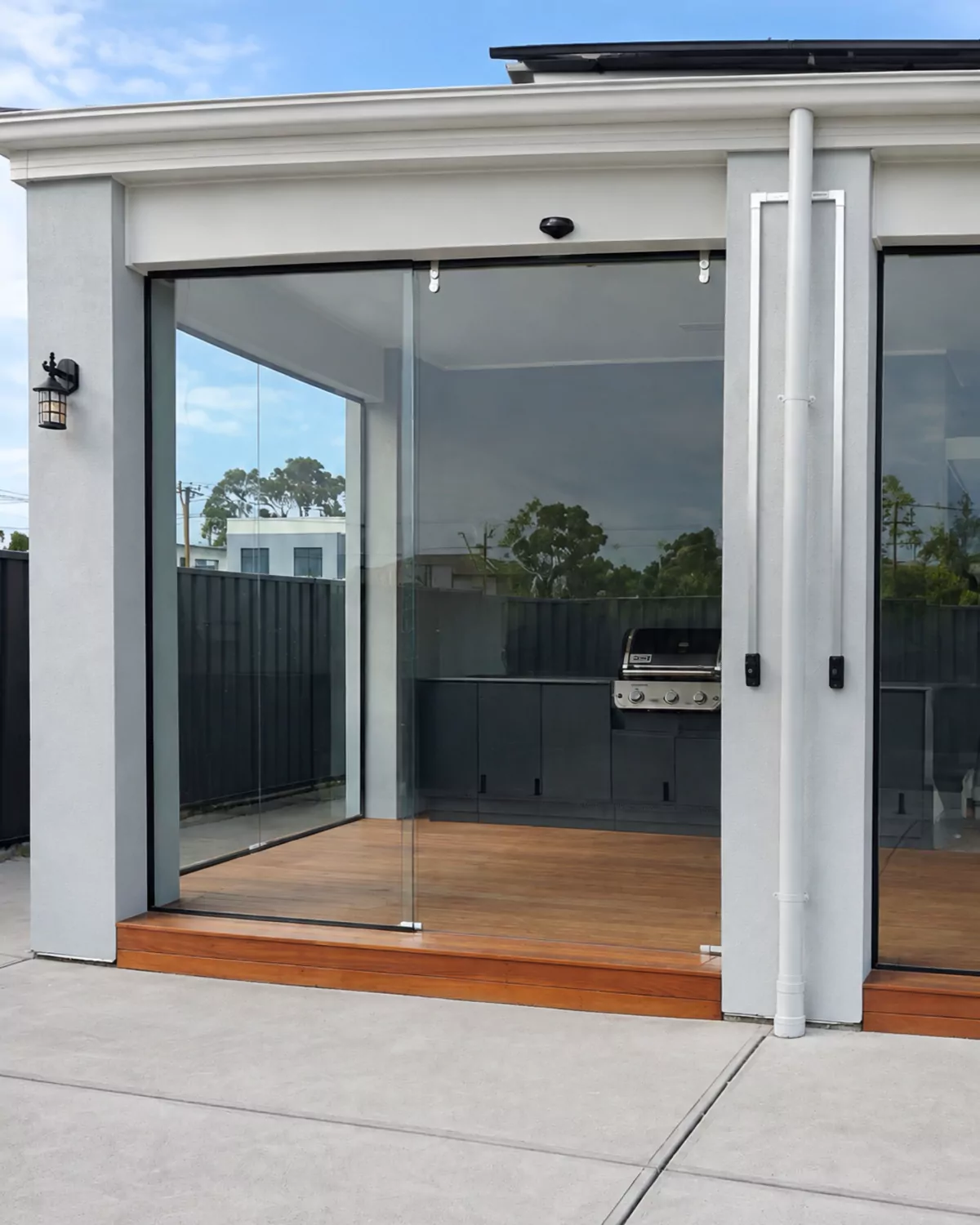 A rendered alfresco on a timber deck enclosed with frameless glass, an outdoor kitchen and barbecue visible inside