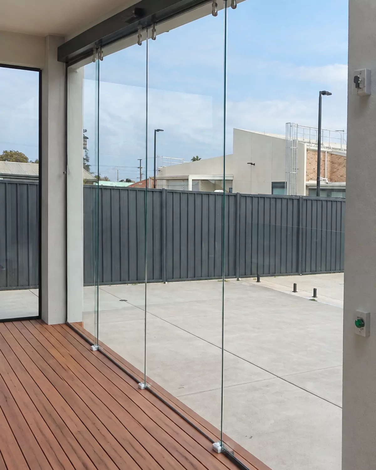 Frameless glass panels hanging from a black top track, slid together at one end of an alfresco, with the timber deck and fence beyond