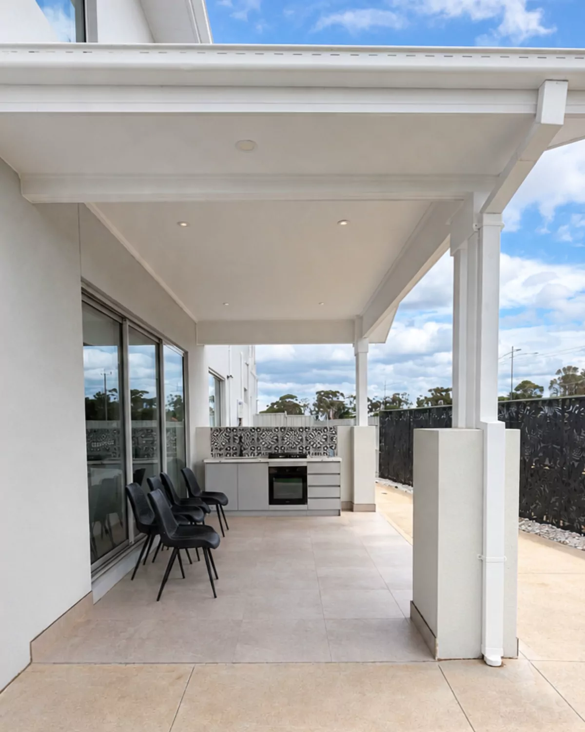 An open, roofed alfresco with an outdoor kitchen, white posts and a tiled floor, before glass was added