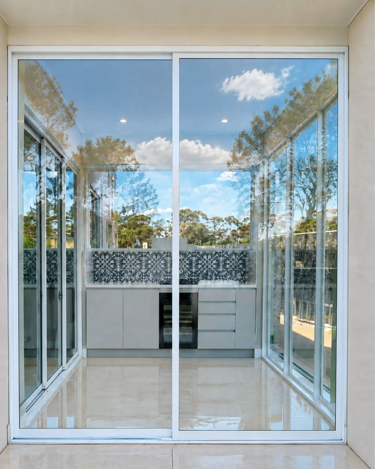 Looking into the finished alfresco through white-framed sliding glass doors, with the outdoor kitchen and patterned splashback inside