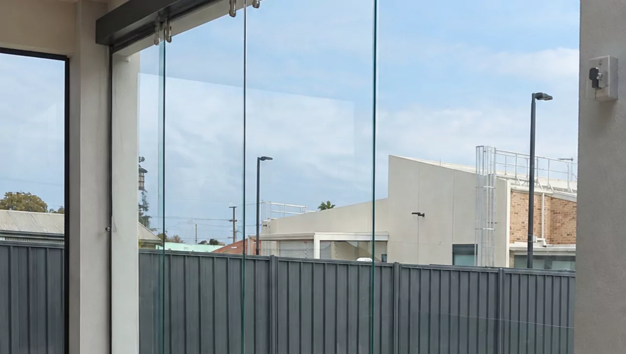 Frameless glass panels hanging from a black top track, slid together at one end of an enclosed alfresco on a timber deck