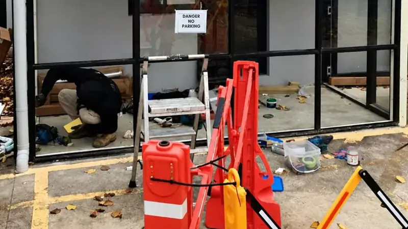 A damaged glass wall with orange barriers in front and a glazier working inside.